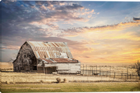 A rustic weathered barn with a corrugated metal roof stands in a dry grass field under a dramatic cloudy sky at sunset, with a fence running along the right side.  All That Remains Giclee canvas gallery wrapped art prints by Teri James